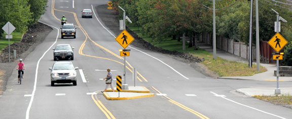 Photo of a pedestrian crossing improvement, woman crossing street with bicyclist using bike lane and cars driving forward yielding to pedestrian